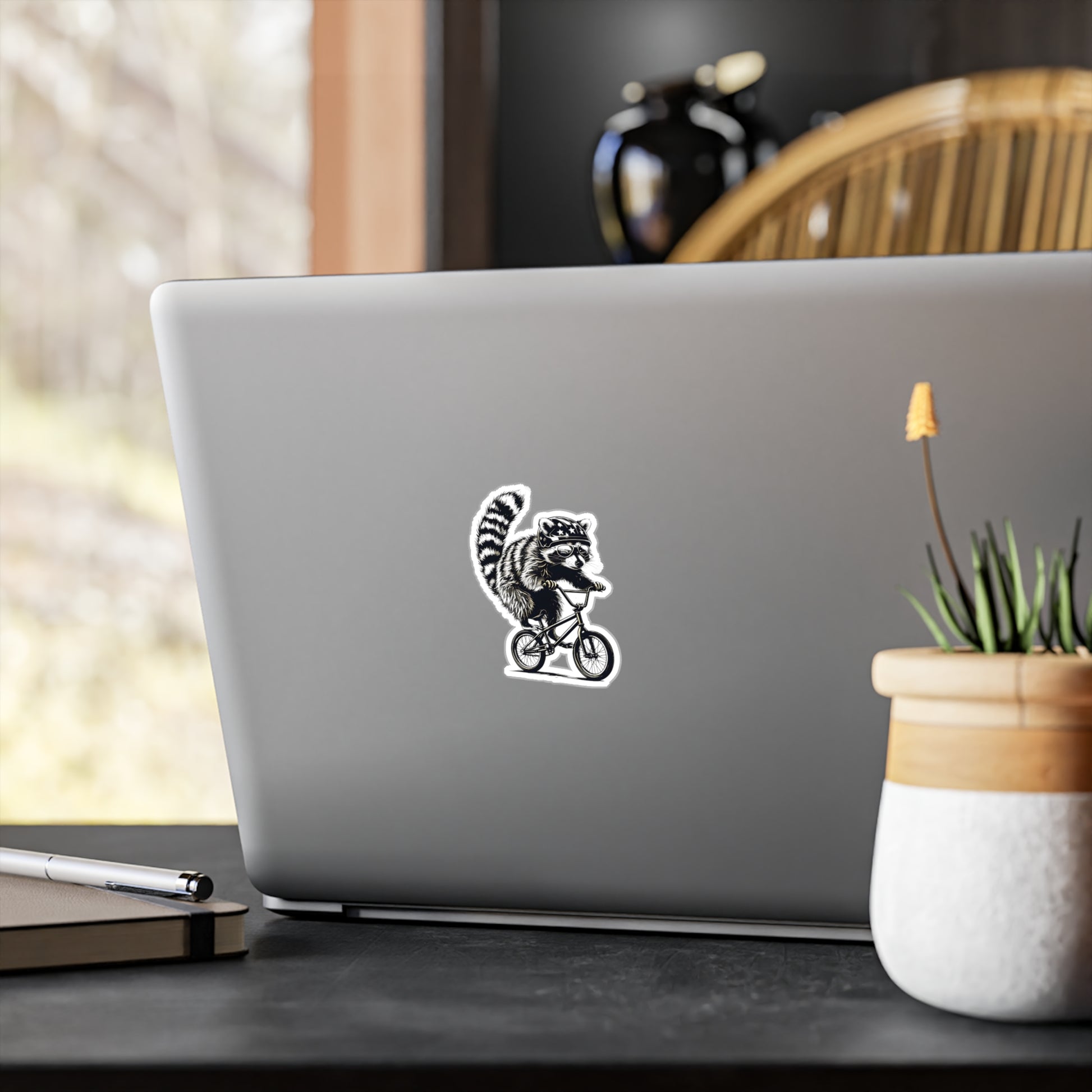 Laptop with a sticker of a raccoon on a bicycle on a desk with a plant and pen.