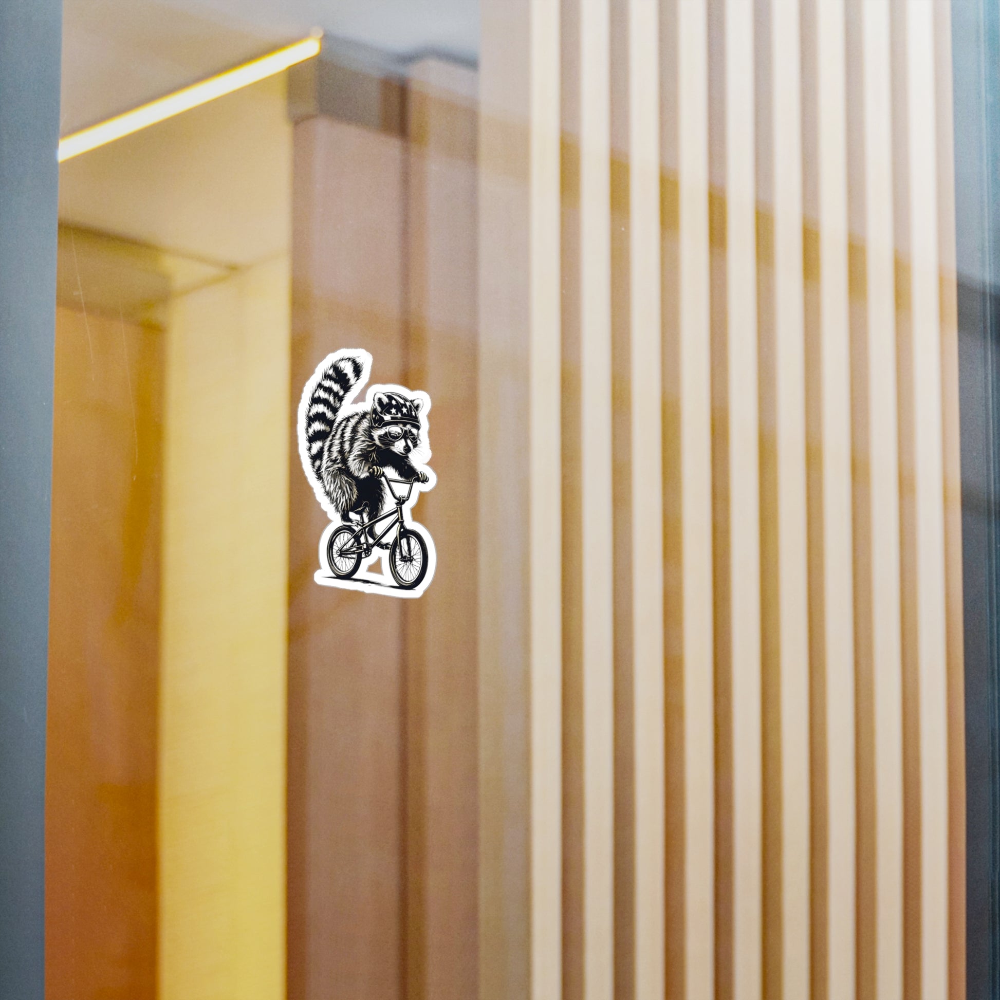 Sticker of a raccoon riding a bicycle on a glass surface with vertical blinds in the background.