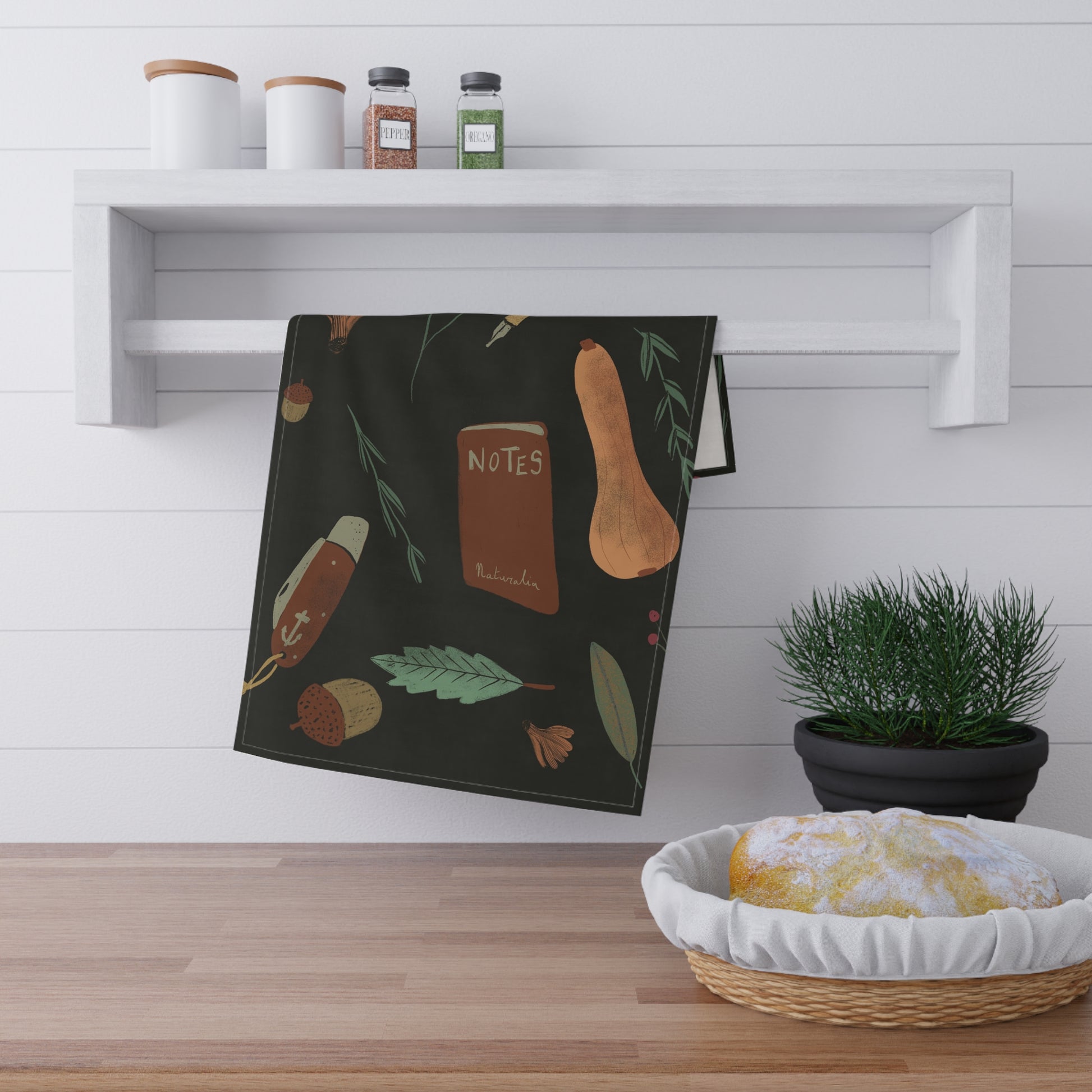 Black towel with autumnal design hanging on a wooden shelf with a plant and basket below.