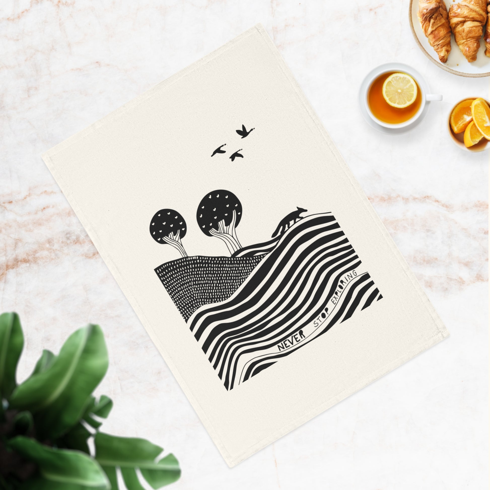 Decorative tea towel with black and white design on a marble surface with a cup of tea and croissants.